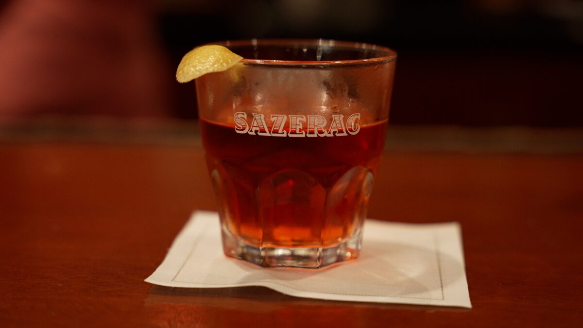 A Sazerac cocktail in a glass printed with the name, garnished with a lemon peel on a bar napkin