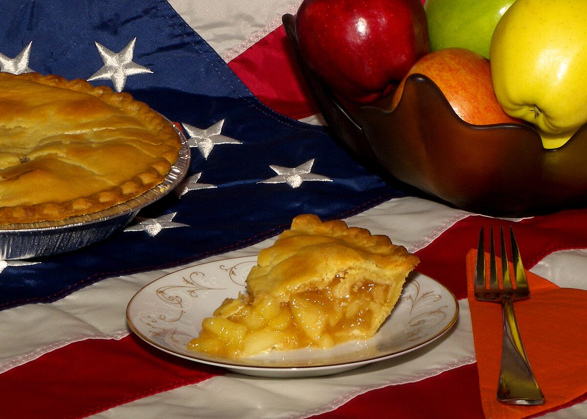 A slice of double-crust apple pie on a plate in front of a whole pie, with apples and an American flag behind