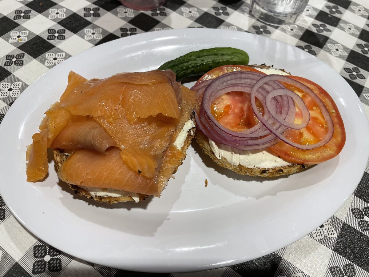 An open bagel topped with cream cheese, smoked salmon, red onion and tomato on a plate with a pickle