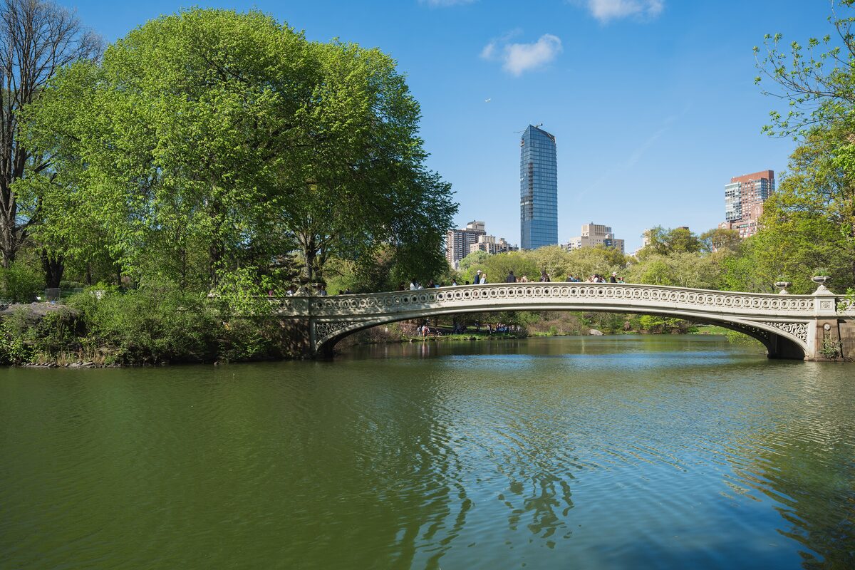 The cast-iron Bow Bridge arching over the Central Park lake in spring, with a tower rising behind the trees