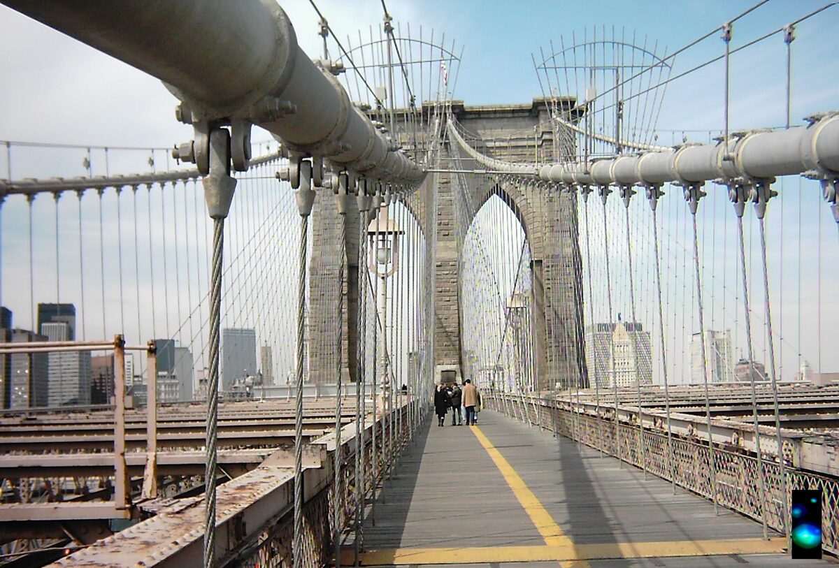 The pedestrian walkway of the Brooklyn Bridge leading toward the stone tower and its pointed arches, with cables fanning overhead