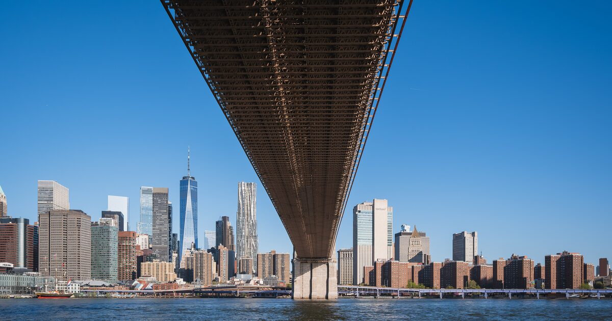 The underside of the Brooklyn Bridge in front of the Lower Manhattan skyline, with One World Trade Center and the Frank Gehry tower under a clear blue sky