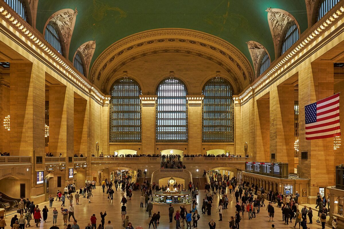 The vaulted Main Concourse of Grand Central Terminal with its tall arched windows, a large flag and crowds crossing the floor