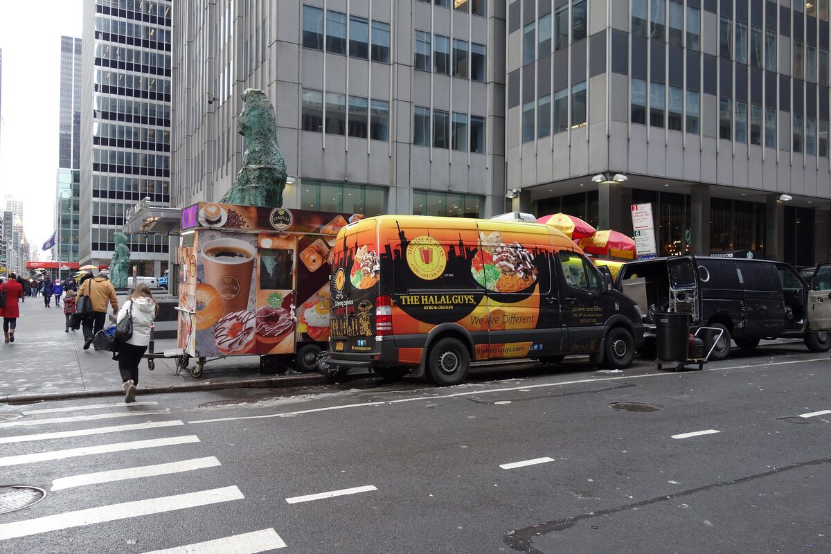 A Halal Guys cart and a bright yellow food van parked on a Midtown street corner under office towers