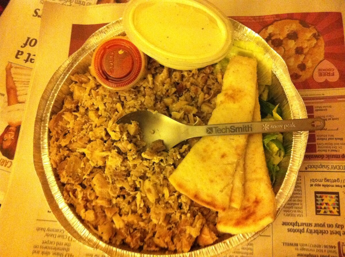 A foil tray of chicken over rice with salad, pita bread and two sauce cups, the classic street cart platter