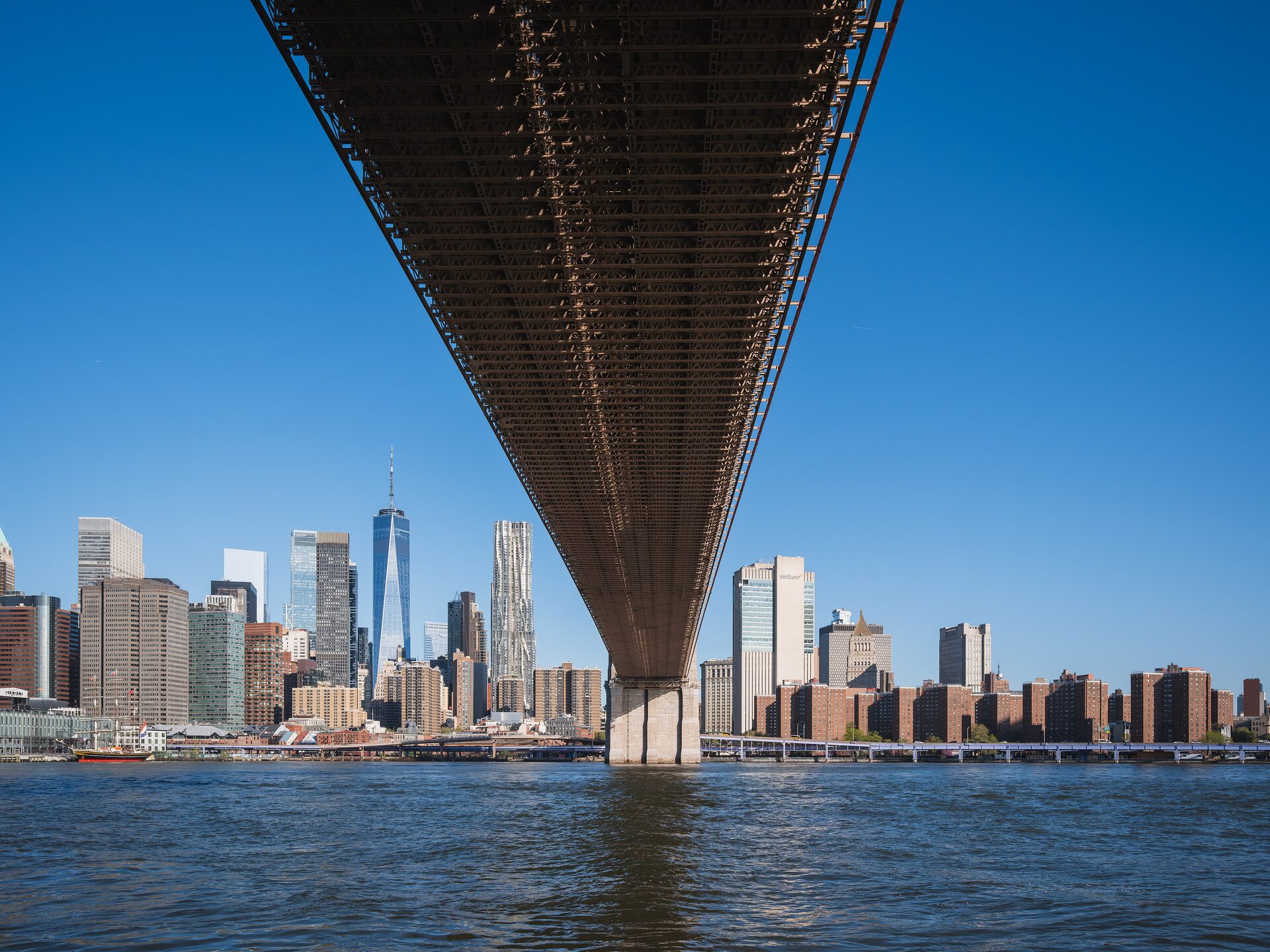 The underside of the Brooklyn Bridge in front of the Lower Manhattan skyline, with One World Trade Center and the Frank Gehry tower under a clear blue sky