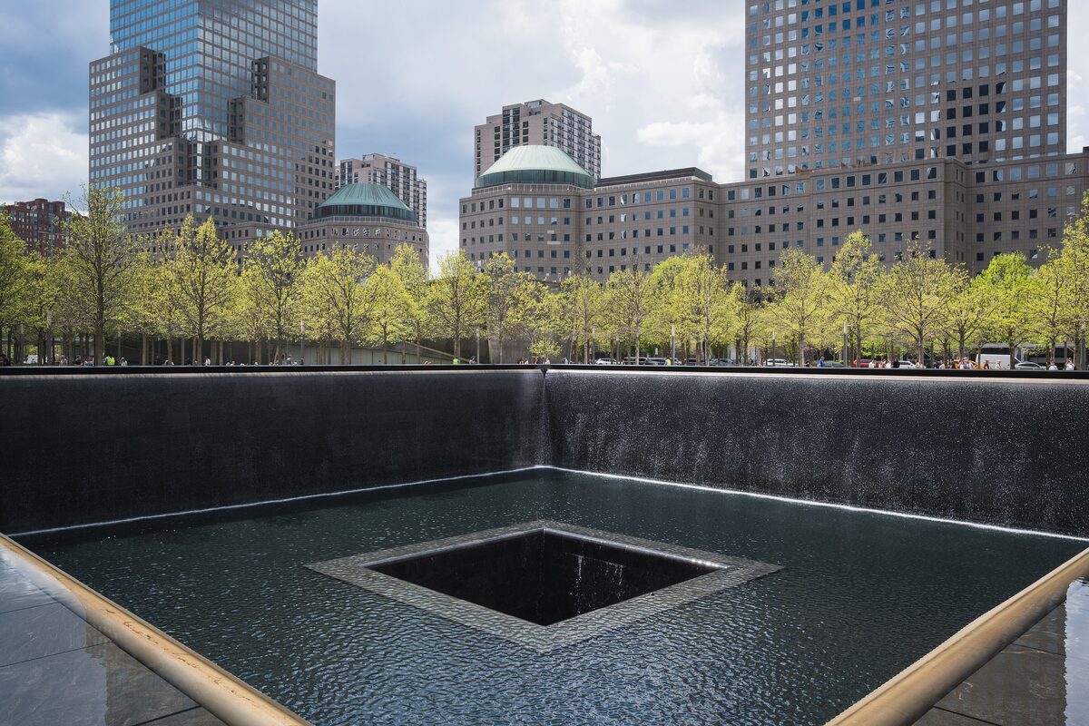 One of the two square reflecting pools of the 9/11 Memorial, with water falling into a central void and trees and towers behind