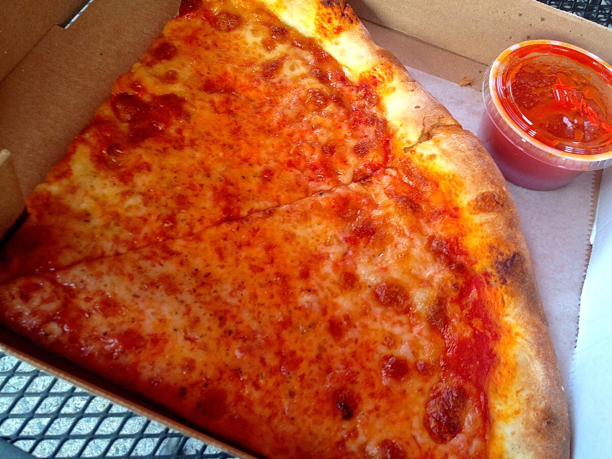 A wide, thin cheese pizza slice on a paper-lined box beside a cup of red sauce