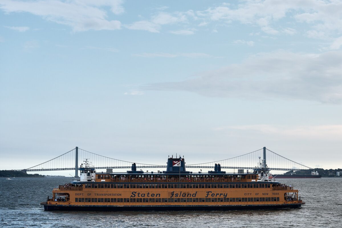 An orange Staten Island Ferry crossing the harbor with the Verrazzano-Narrows Bridge in the distance