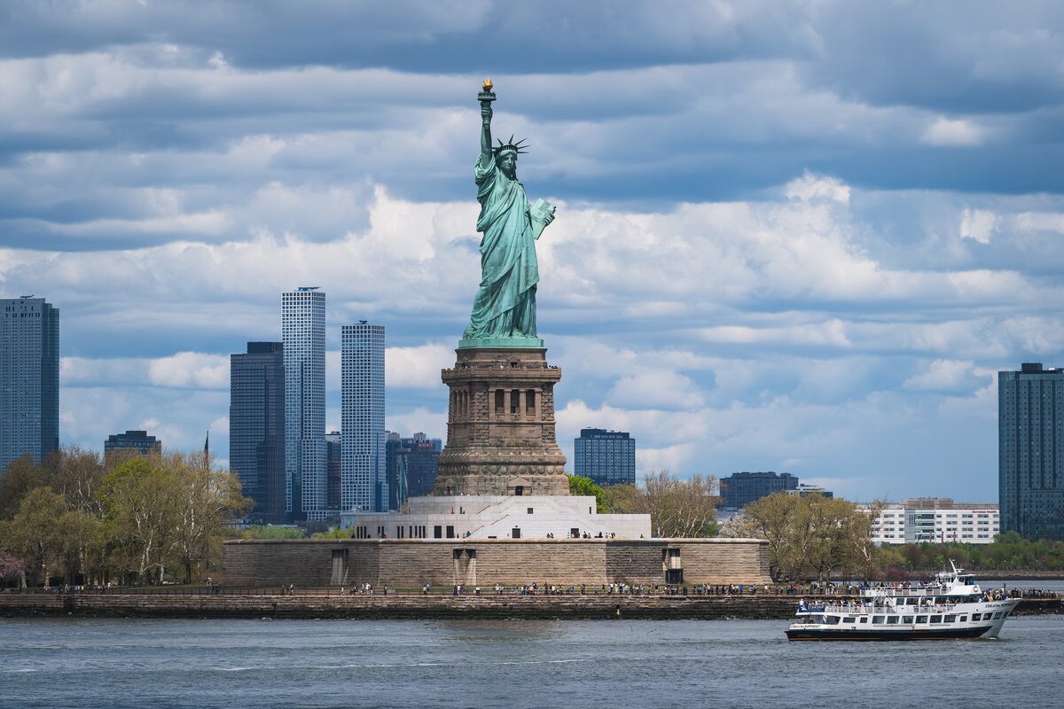 The Statue of Liberty on Liberty Island with a sightseeing boat passing in front and tall buildings behind