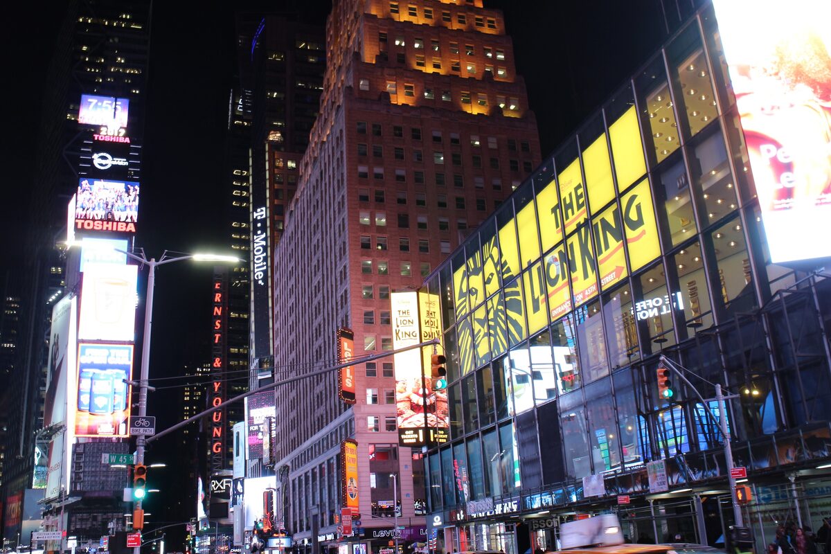 Times Square at night with glowing billboards and the yellow Lion King theater sign on the corner