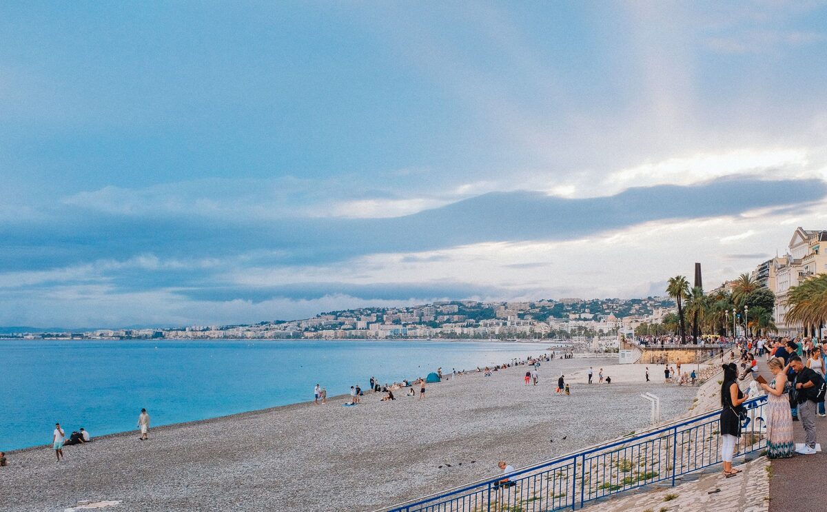 The long gray pebble beach of Nice with swimmers and walkers, the Promenade railing and the coastline curving away