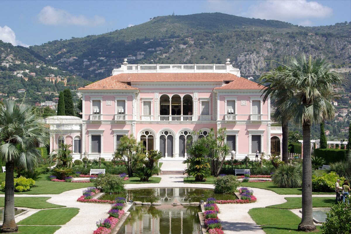 The pink Villa Ephrussi de Rothschild behind its formal French garden and long pool, with green hills behind