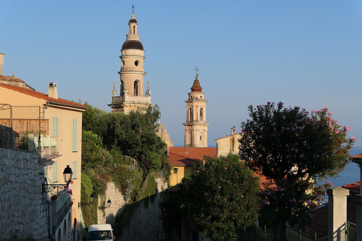 Two Baroque bell towers rising above old houses and trees in Menton, with the sea on the right
