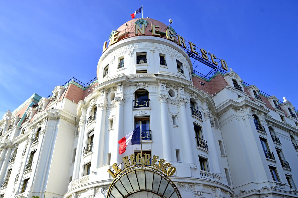 The white Belle Époque facade and dome of the Hotel Negresco with its name in gold letters and flags, under a blue sky