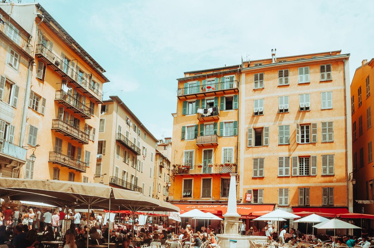 Café terraces with umbrellas on a square in Old Nice, surrounded by tall ocher and yellow houses with green shutters