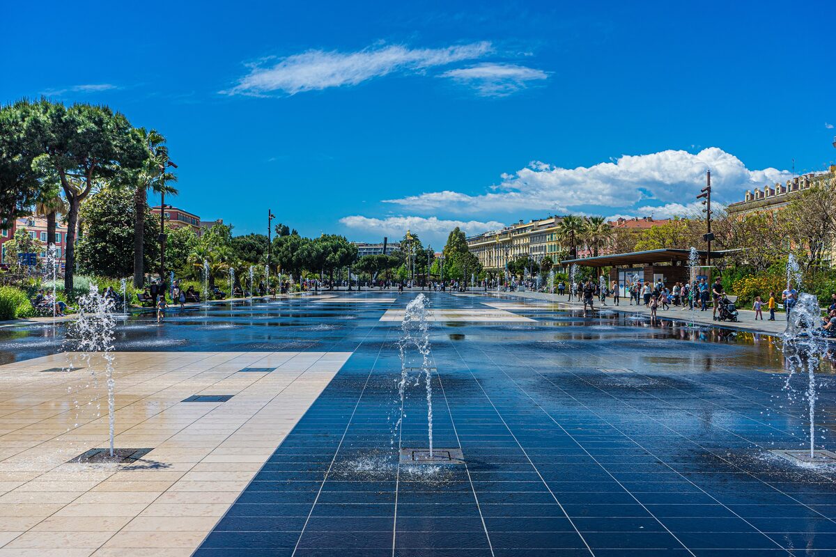 Fountain jets rising from the shallow water mirror of the Promenade du Paillon, with palm trees and buildings behind