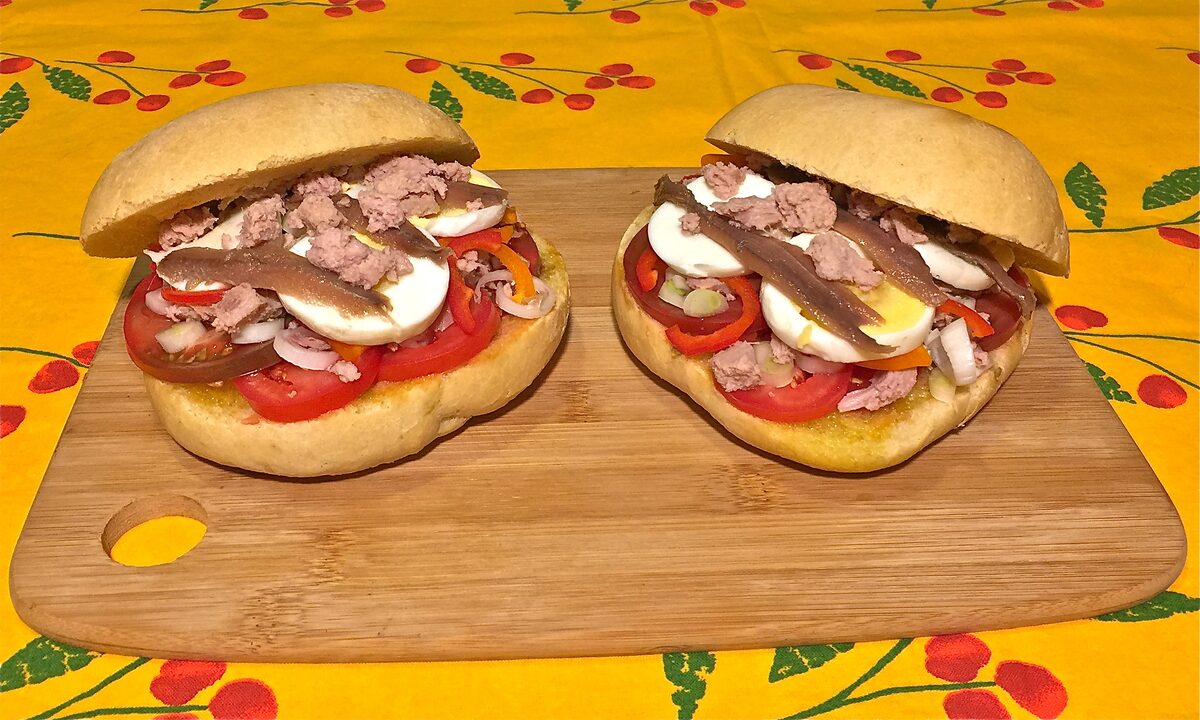 Two round pan bagnat sandwiches filled with tuna, egg, tomato and anchovies on a wooden board