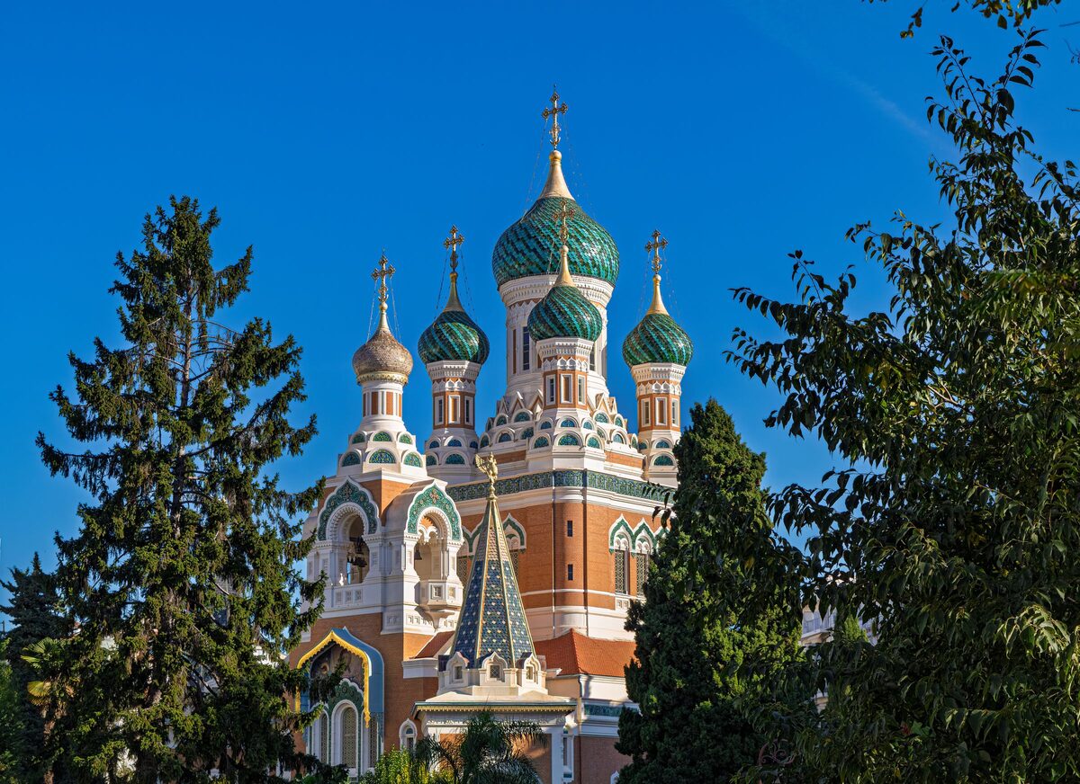 The green and gold onion domes and pink and white facade of the St. Nicholas Russian Orthodox Cathedral in Nice, framed by trees against a blue sky
