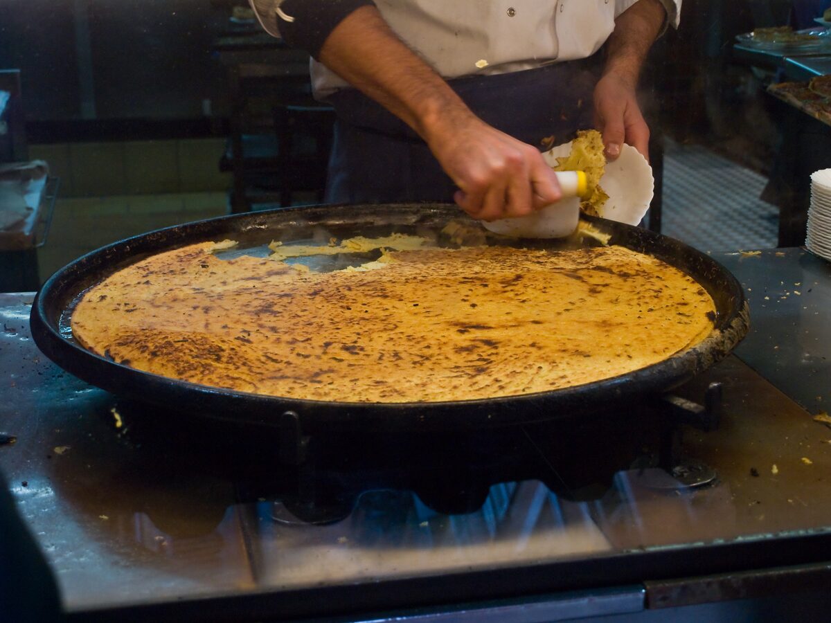 A cook spreading socca in a huge round black pan, the chickpea pancake golden and blistered