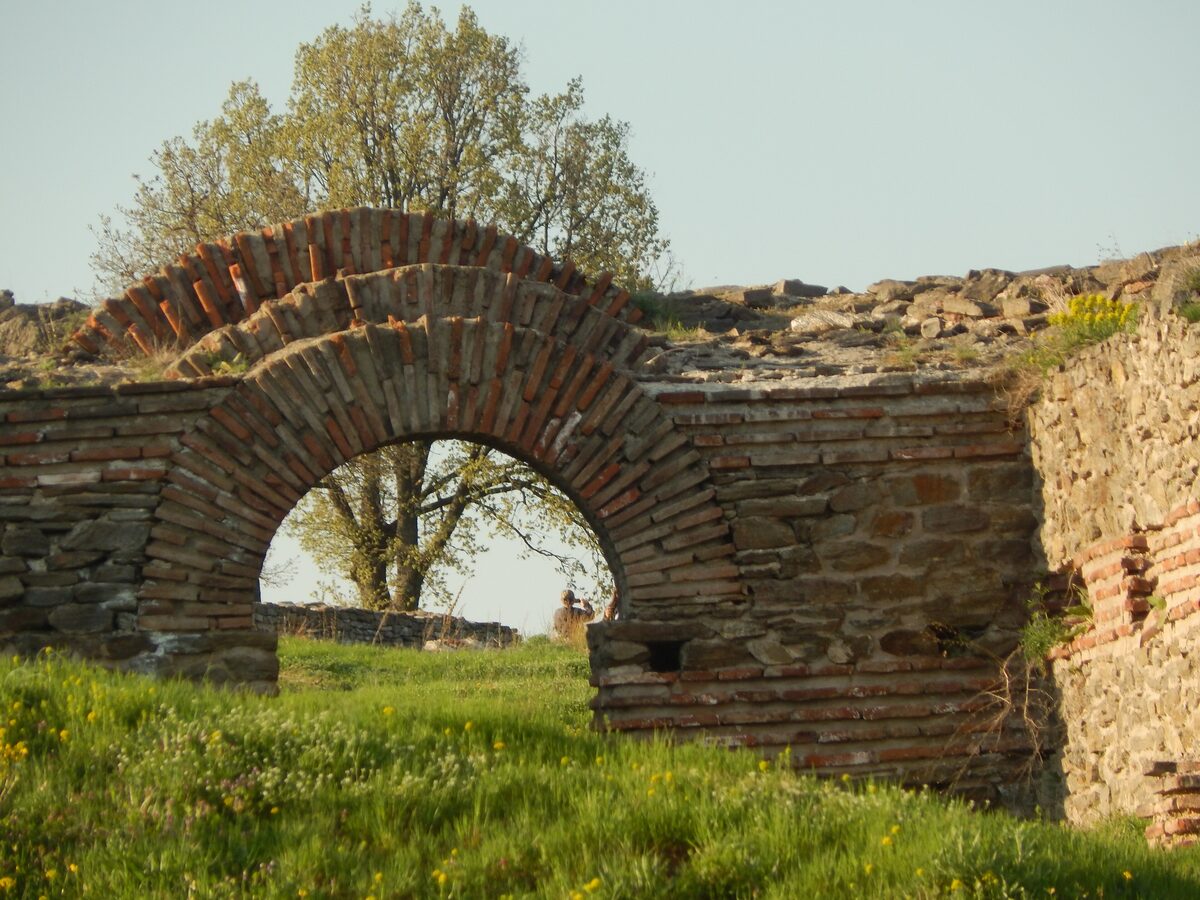 A layered brick and stone arch among the grassy ruins of Caričin Grad (Justiniana Prima), lit by low evening sun with a tree behind.