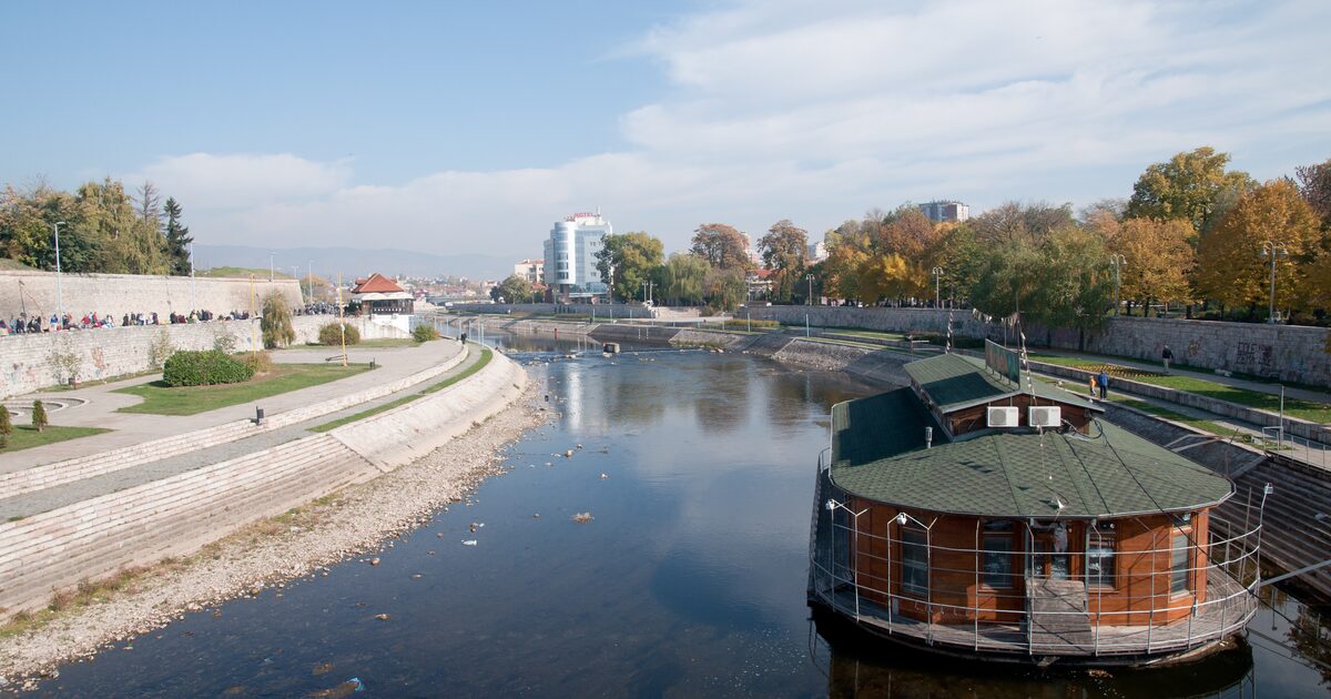 The Nišava river flowing through Niš on an autumn day, with the fortress walls and quay on the left, a floating river café on the right and the city behind.