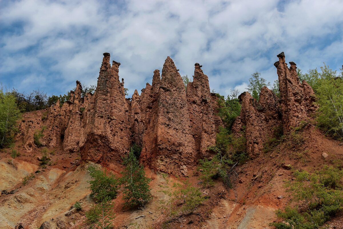 The reddish earth pillars of Đavolja varoš, some still capped with stone, rising from an eroded slope under a cloudy blue sky.