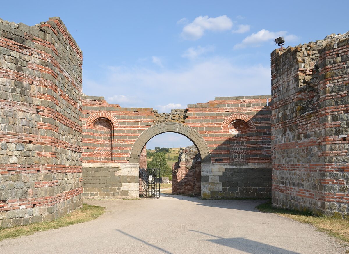 The arched gateway of Felix Romuliana at Gamzigrad, framed by massive walls of alternating stone and red brick bands under a blue sky.