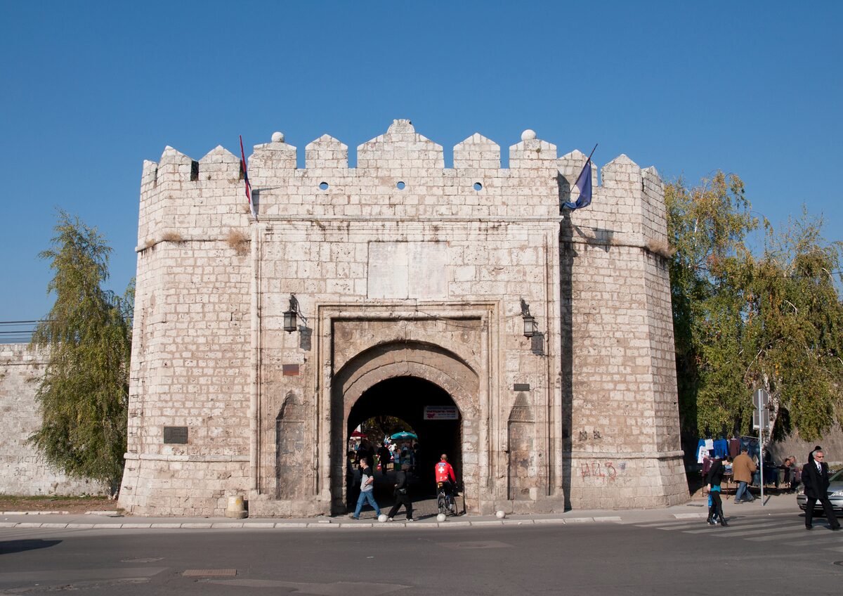 The crenellated stone Stambol Gate of Niš Fortress with its arched entrance under a clear blue sky, people walking through.