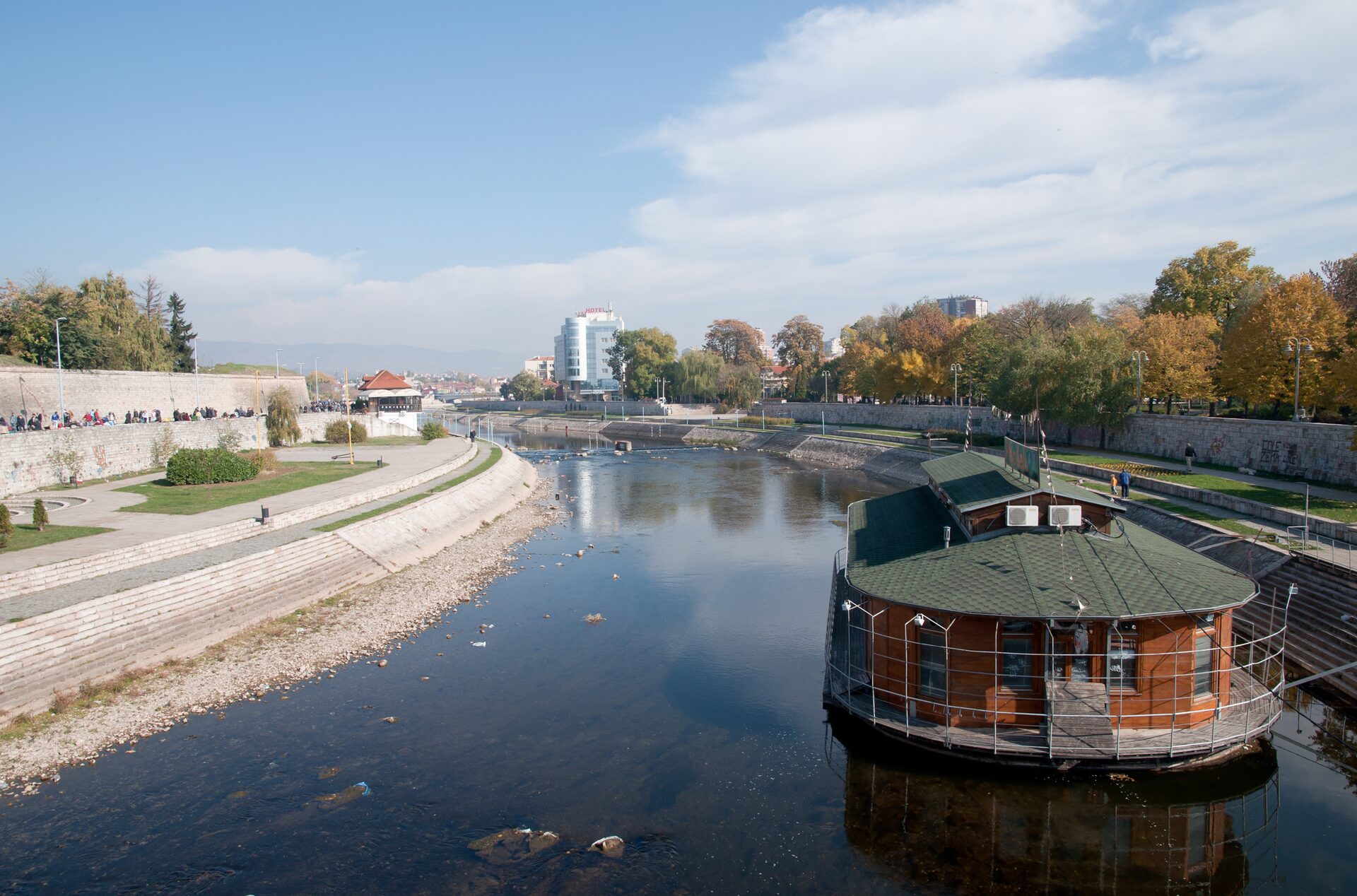 The Nišava river flowing through Niš on an autumn day, with the fortress walls and quay on the left, a floating river café on the right and the city behind.