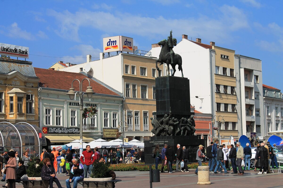 The equestrian Monument to the Liberators of Niš on King Milan Square, with old facades, café umbrellas and people strolling around it.