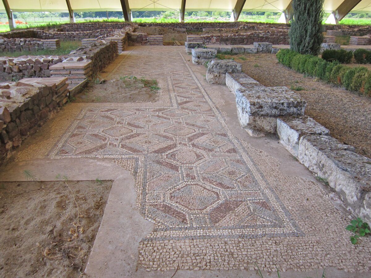 A geometric Roman floor mosaic running between low stone walls of the villa at Mediana, under the site's protective roof.