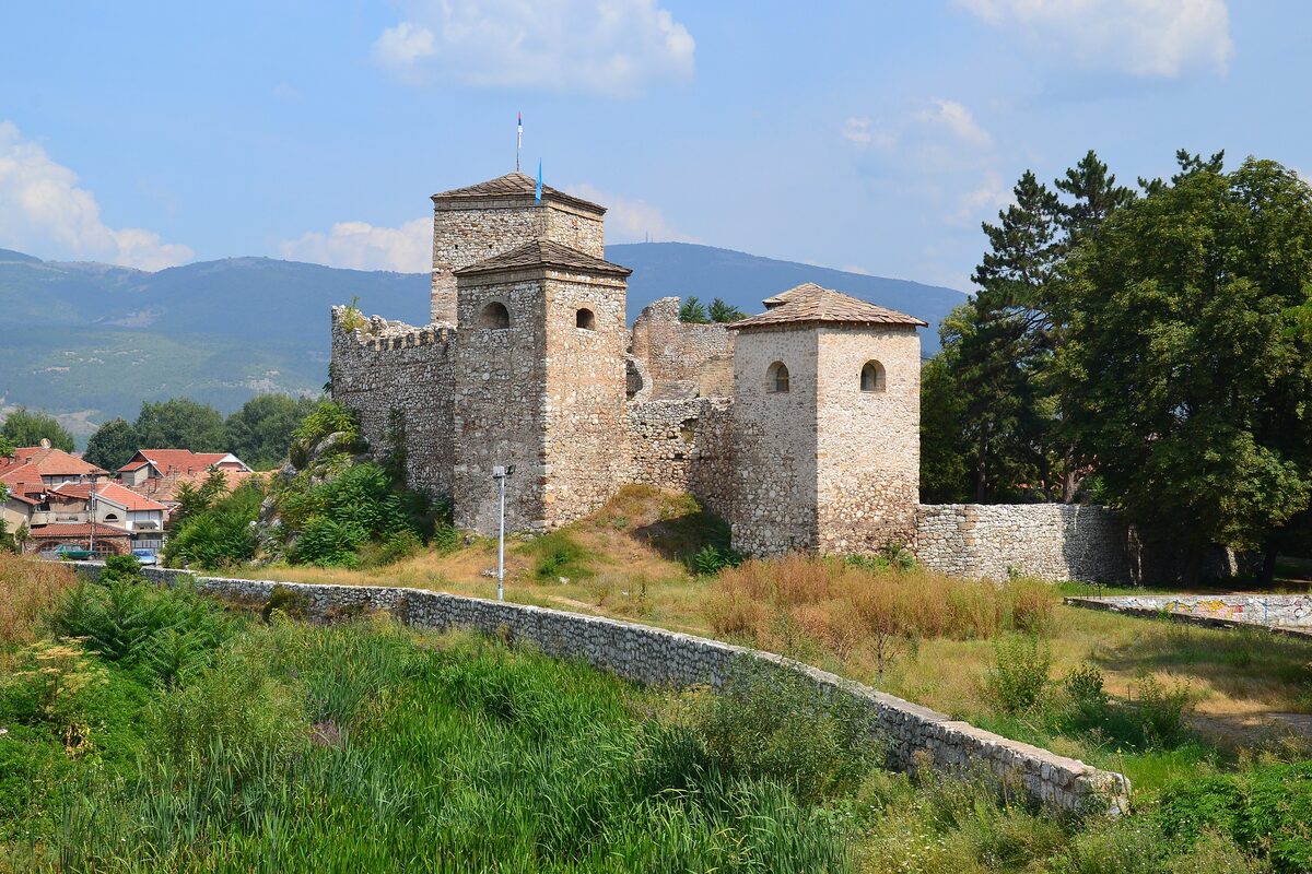 The stone towers and walls of Pirot's Kale fortress (Momčilov grad) above a grassy bank, with red roofs and mountains behind.