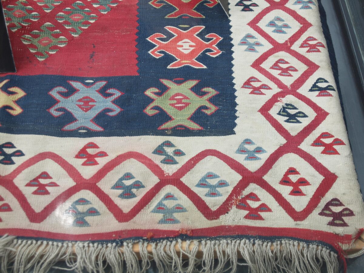 The corner of a fringed Pirot kilim in red, navy and white, woven with geometric diamond borders and stylised motifs, displayed in a museum case.
