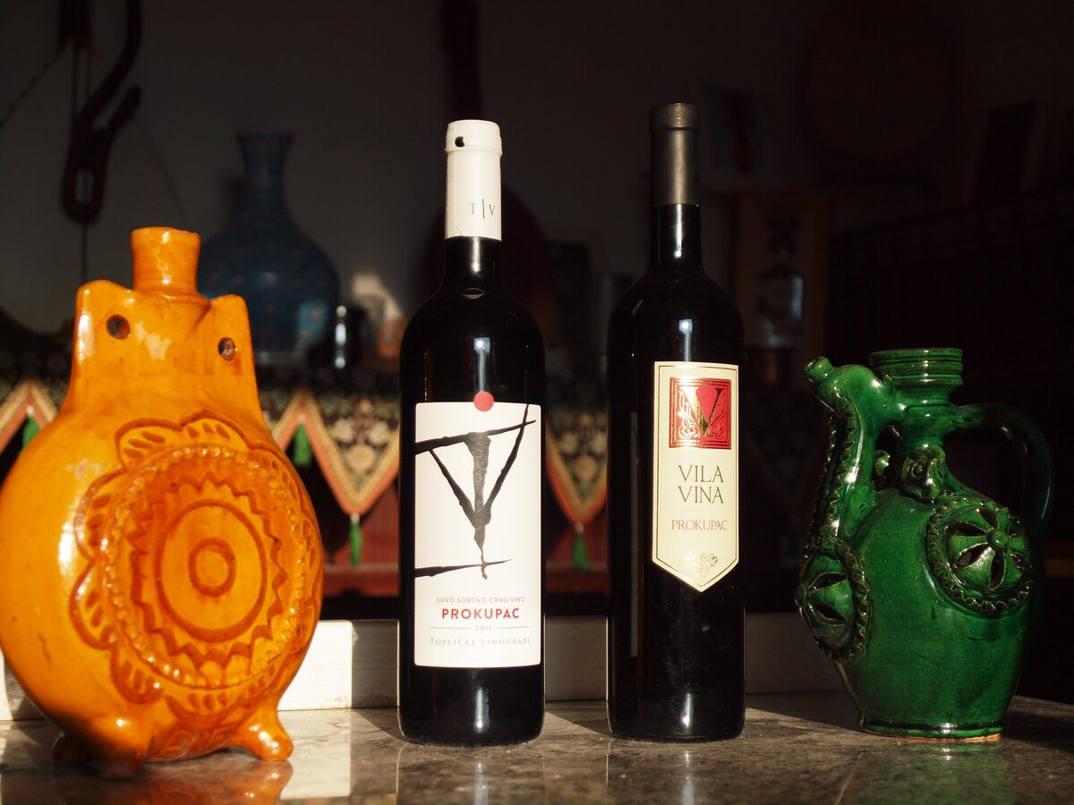 Two bottles of Serbian Prokupac red wine, one from Toplički vinogradi near Prokuplje, between a glazed orange flask and a green ceramic jug.