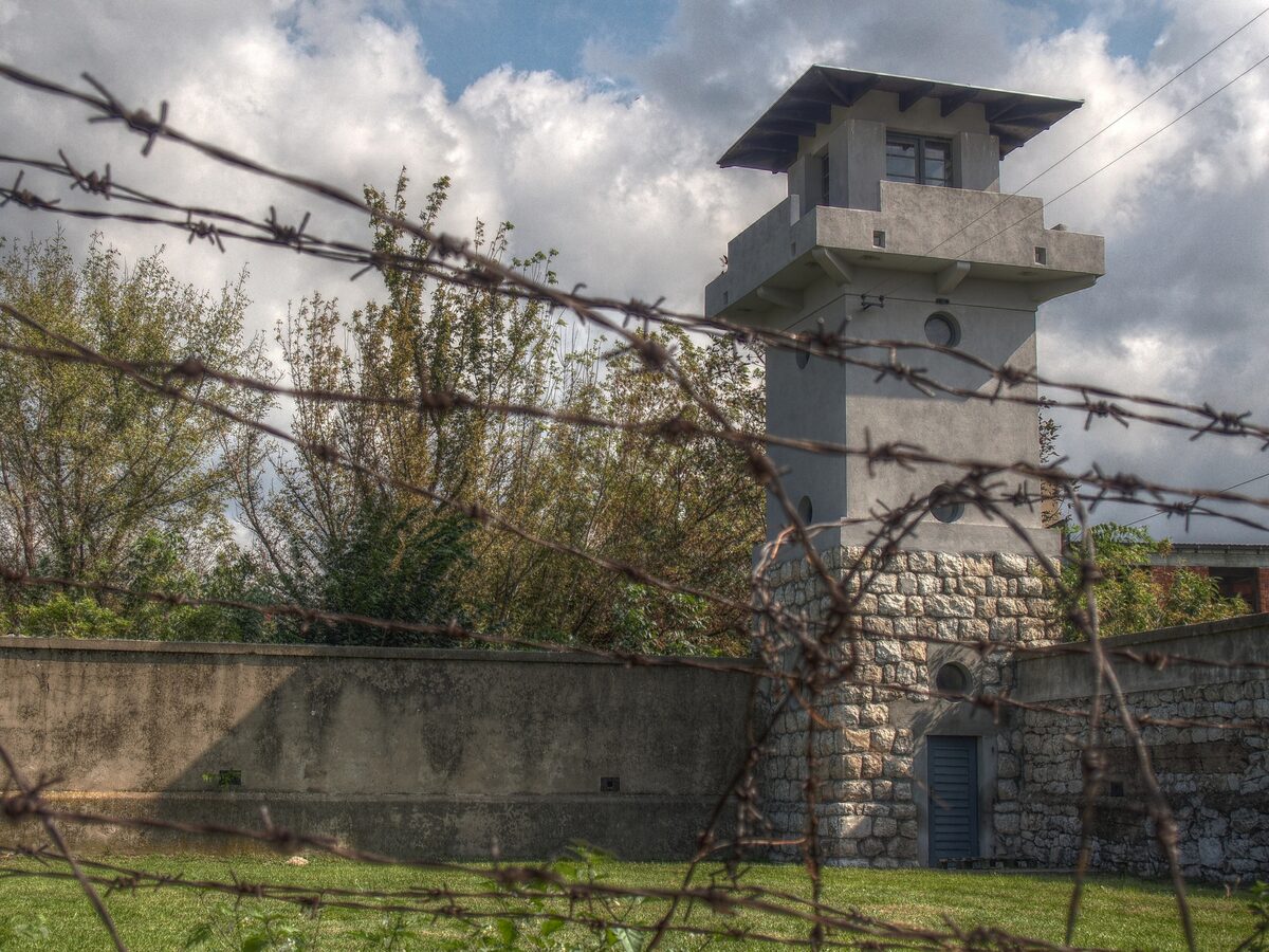 A guard tower and the perimeter wall of the Crveni Krst camp, seen through strands of barbed wire under a cloudy sky.