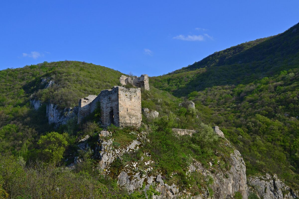 The ruined stone towers of Sokograd fortress on a rocky outcrop above the Moravica gorge, surrounded by green wooded hills.