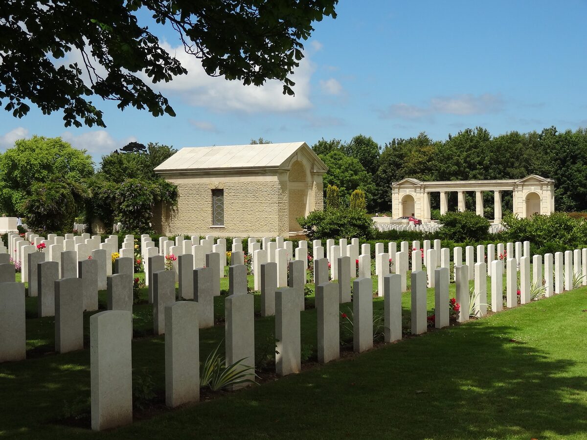 Rows of white headstones on the lawn of the Bayeux War Cemetery, with the colonnade of the Bayeux Memorial in the distance