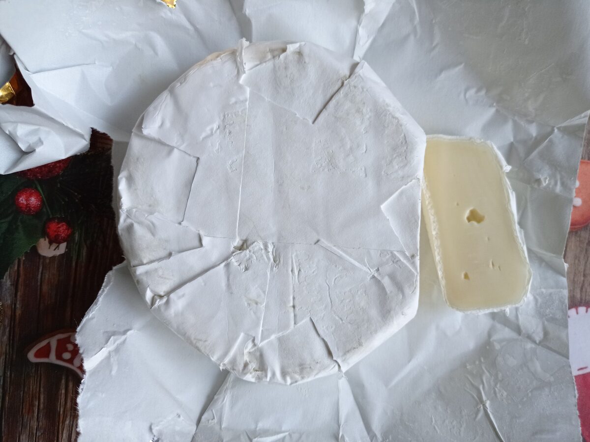 A round Camembert in its white paper wrapper, with one wedge cut off to show the pale, soft inside