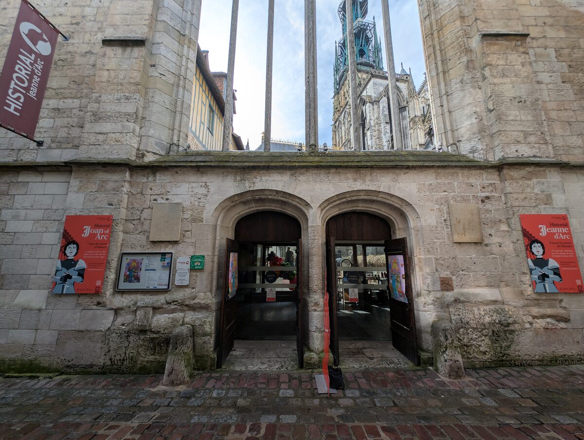 The entrance of the Historial Jeanne d'Arc in a stone wall, with the spire of the cathedral visible above it