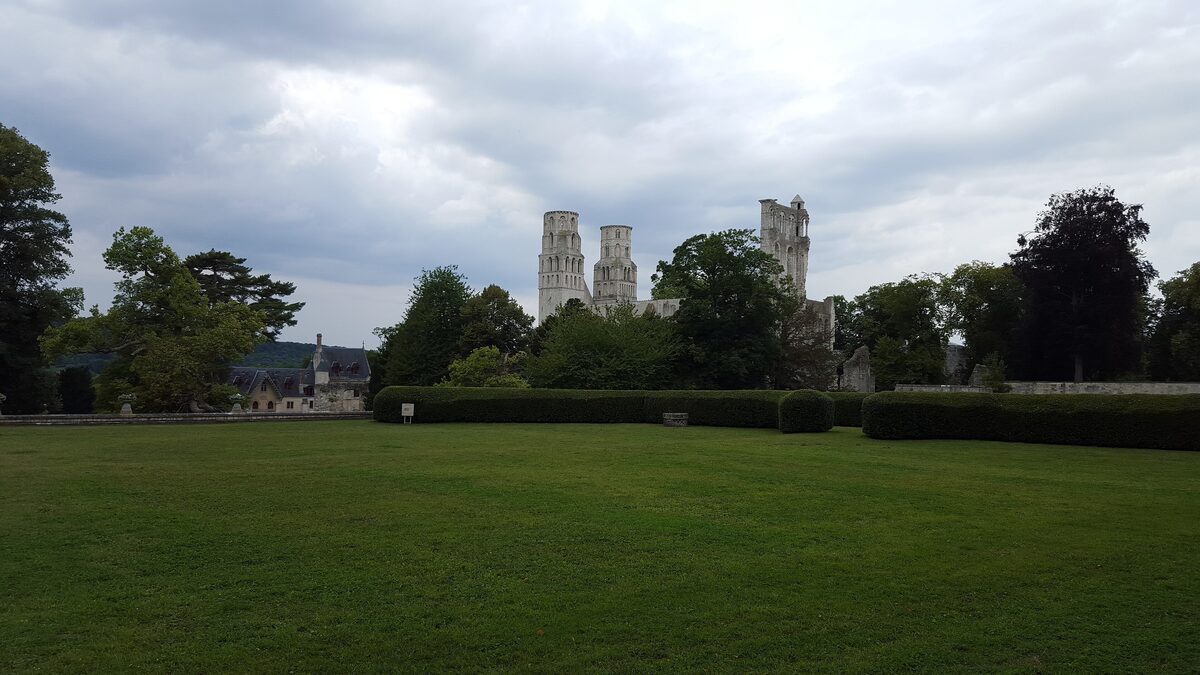The ruined Jumièges Abbey towers rising behind a lawn and a clipped hedge, with an old stone house on the left
