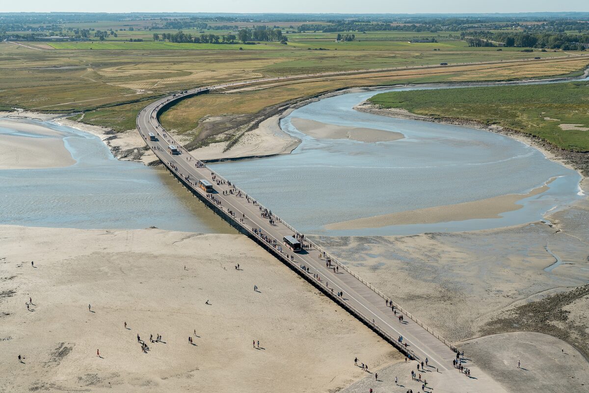 Aerial view of the causeway to Mont-Saint-Michel, with walkers and shuttle buses crossing the sand and the water channels