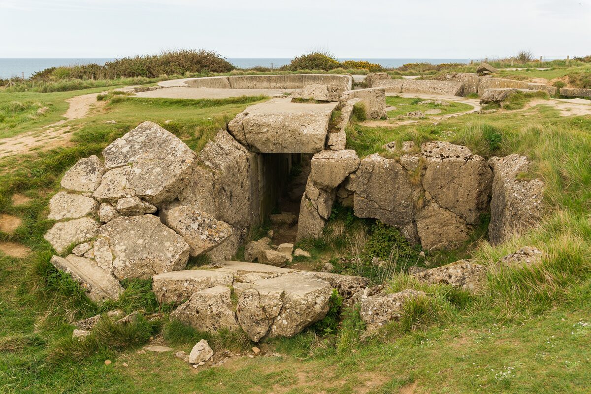 A concrete German gun bunker at Pointe du Hoc, its roof split and the broken blocks tumbled over the grass