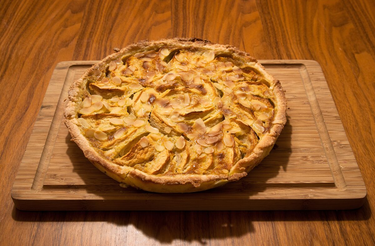 A round Norman apple tart with thin apple slices and flaked almonds on top, on a wooden board
