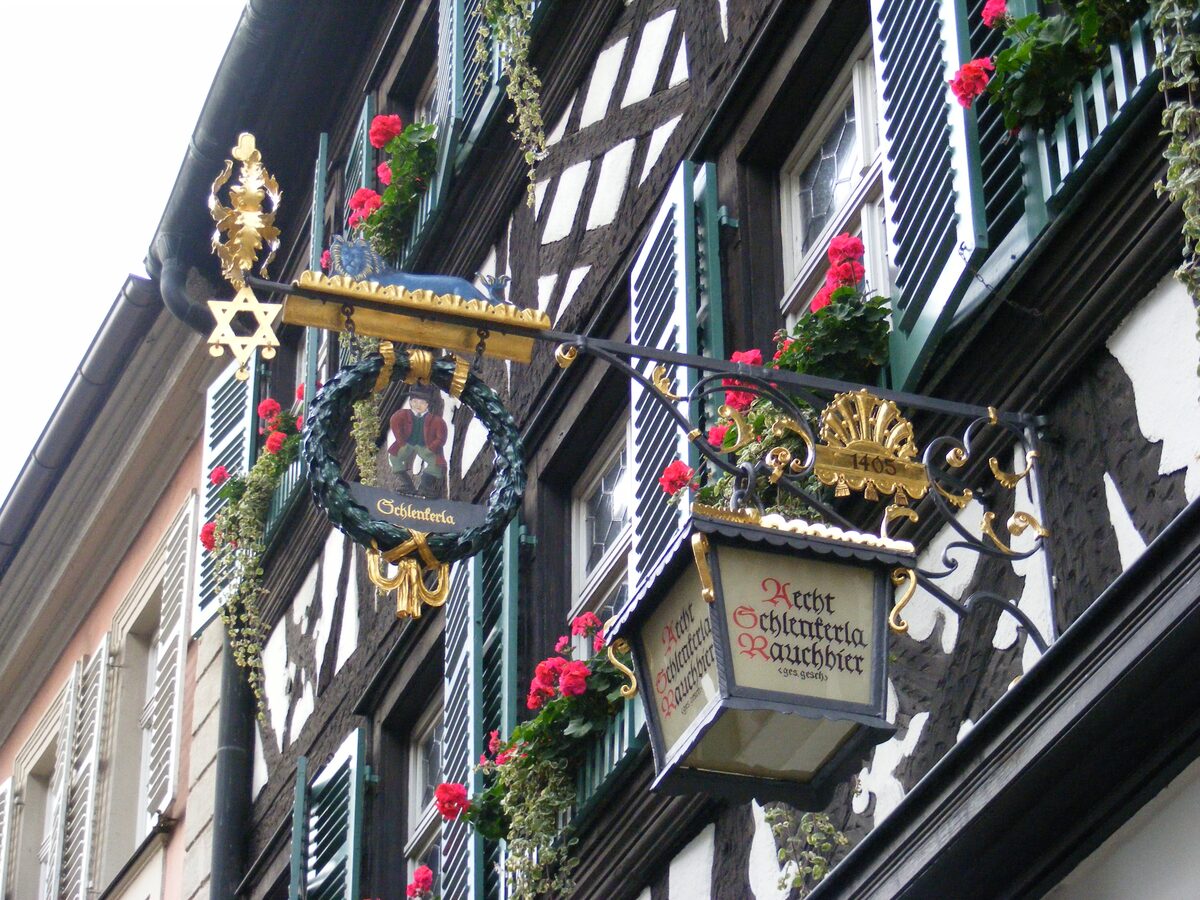 The hanging sign of the Schlenkerla tavern with a gilt frame, set on a half-timbered facade with red flowers