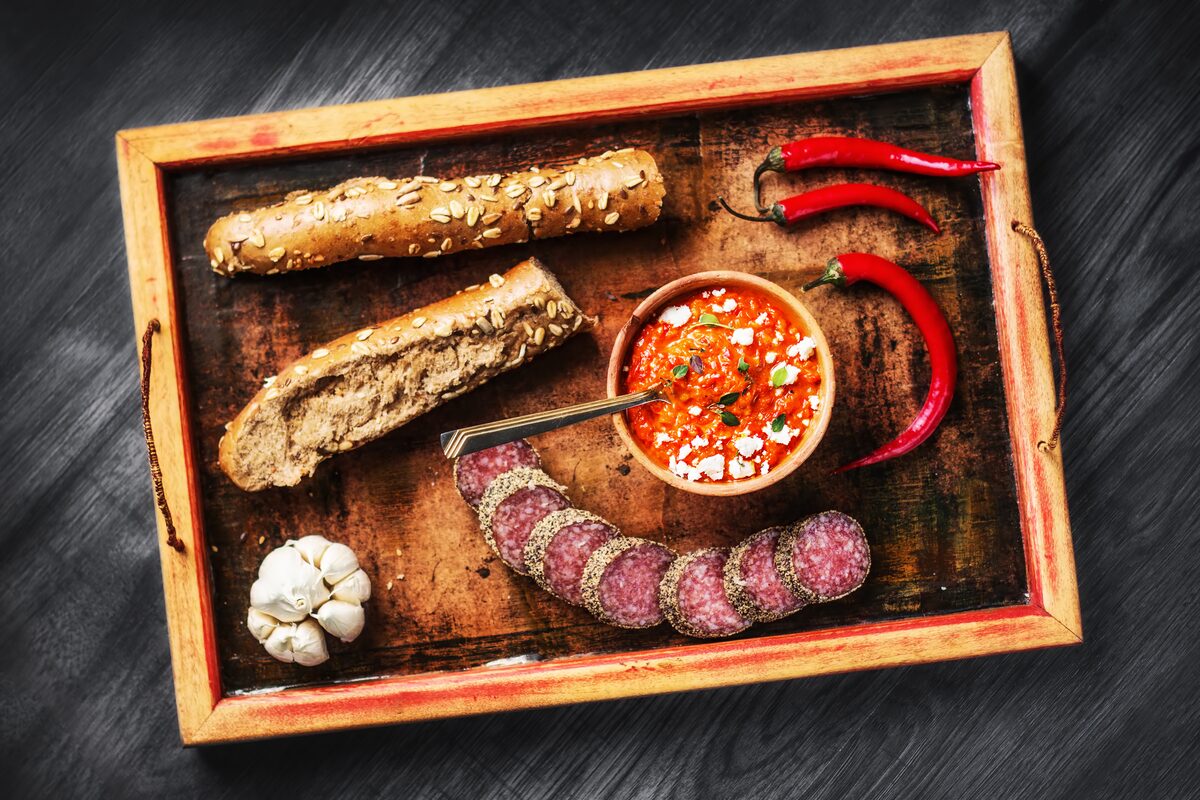 Ajvar, the red pepper spread, in a bowl on a tray beside bread, sausage and chili