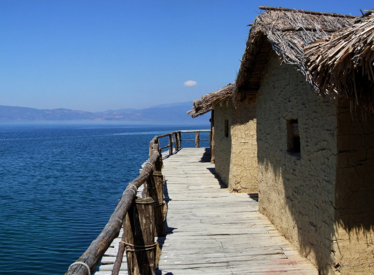 A wooden walkway on stilts leading past a reed-roofed clay house of the Museum on Water, with the lake on the left