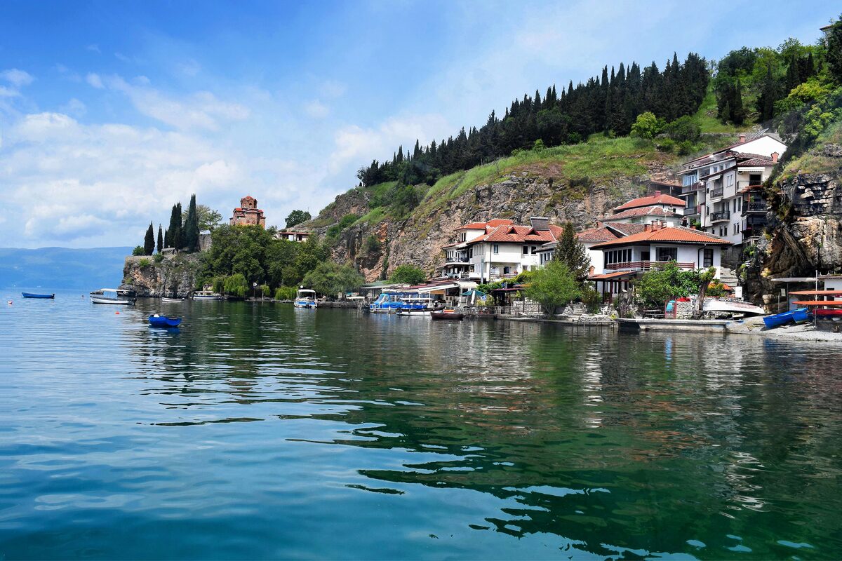 The Kaneo shore with red-roofed houses under a wooded slope and the church on the point, over calm blue-green water
