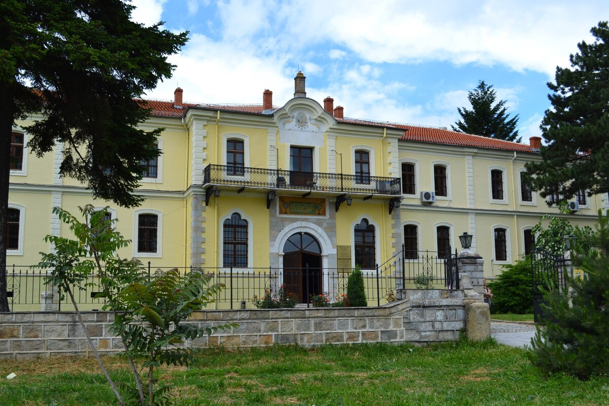 The yellow neoclassical building of the Museum of Bitola with red roofs, behind a stone wall and pine trees