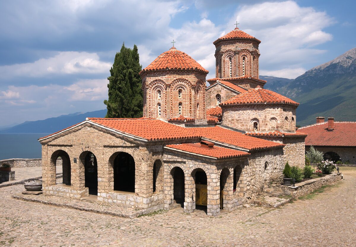 The stone church of St. Naum with its two red-tiled domes and an open arcade, with the lake and mountains behind